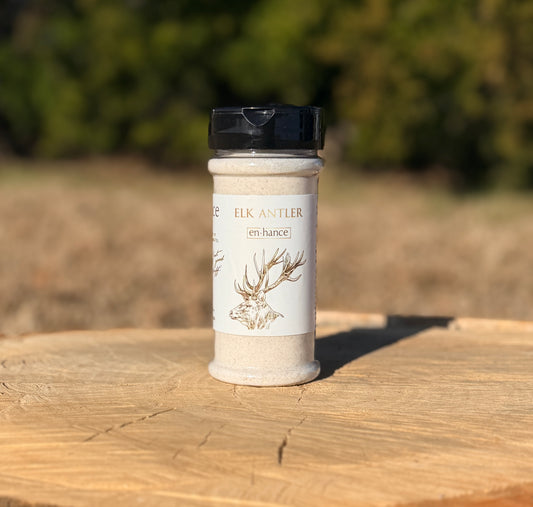 A bottle of Elk Antler seasoning stands on a rustic wooden surface with a blurred outdoor forest background. The product features a black cap, a white label with a brown elk antler illustration, and light beige seasoning inside.